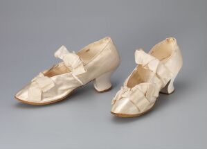  A pair of antique women's cream-colored satin shoes with woven front detail and large bows, displayed side by side on a gray background.