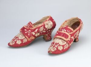  A pair of ornate red fabric women's shoes with cream floral patterns, pointed toes, and decorative metallic buckles on a neutral gray background.