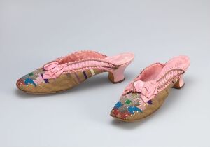  A pair of vintage beige women's flat shoes with small heels, featuring intricate floral embroidery in shades of blue, pink, green and yellow, embellished with light pink ruffled trim around the openings. The shoes are displayed against a plain, light-colored background.