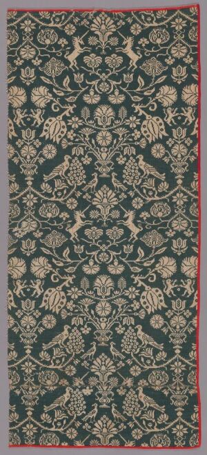  An elaborate forest green textile or rug with intricate golden, white, and lighter green floral-like patterns, bordered on one side by a narrow salmon pink strip, suggesting traditional craftsmanship and design.