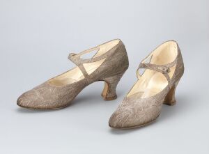  A pair of vintage women's T-strap shoes in a neutral beige color, with medium heels and rounded toes, displayed against a plain background. Artist name and title are unknown.