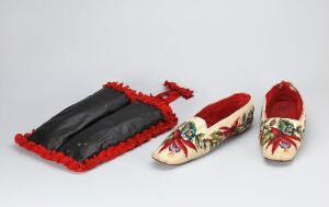  A pair of traditional cream-colored shoes with red, green, and yellow floral embroidery next to a black pouch with red fringe and a drawstring.