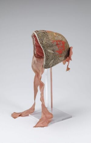  An eerie artistic piece with a patterned hood-like garment showing red, green, and beige floral motifs, from which two realistic human legs emerge, set against a plain gray backdrop. The legs and garment are displayed on a stand, and the detail in the legs’ skin suggests a lifelike quality, whereas the garment hints at historical clothing. Artist name and title are unknown.