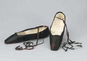  A pair of gently used black ballet pointe shoes with long, black satin ribbons lying on a light gray background, showing signs of wear distinctive to dancers' footwear.