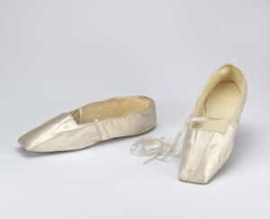  A pair of pale cream-colored ballet shoes with satin finish and untied ribbons, displayed on a neutral grey background. They appear new and unused, capturing the elegance of classical ballet dancewear.