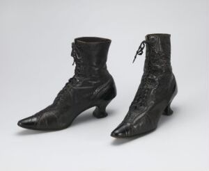  A pair of black, glossy vintage high-heeled lace-up boots with elaborate designs on the vamp, set against a neutral light grey background.