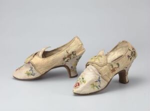  A pair of vintage cream-colored women's shoes with low heels, adorned with a subtle floral pattern in pastel shades, a pointed toe, and decorative bows, set against a neutral grey background.