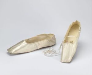  A pair of pale ivory ballet slippers made of a satin-like material, one lying flat and the other standing upright, against a subtle light grey background.