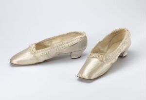  A pair of vintage, ivory-colored silk or satin women's shoes with a low heel and ruched detail around the openings, set against a light grey background.