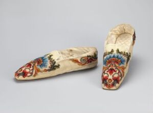  A pair of cream-colored, pointed-toe women's slippers with red, blue, and green floral embroidery, placed against a neutral grey background.