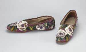  A pair of vintage flat shoes with a royal purple fabric upper, beautifully embroidered with white and pink flowers and green leaves, displayed against a neutral gray background.