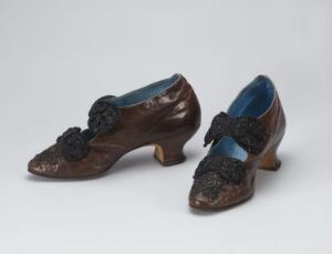  A pair of dark brown vintage women's shoes with modest heels and black floral embellishments on the toes, displayed against a light grey background. The interior is lined with a striking blue material, and the shoes exhibit a polished finish with signs of gentle wear.