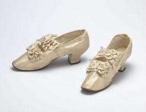  A pair of elegant vintage off-white women's shoes with decorative floral embellishments on the vamps, set against a plain light grey background.