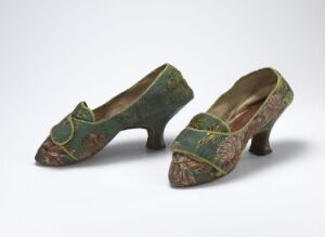  A pair of ornate antique women's shoes with curving medium heels, featuring a moss green brocade fabric with a floral pattern accented in red, gold, and pink, highlighted by gold stitching, against a neutral background.