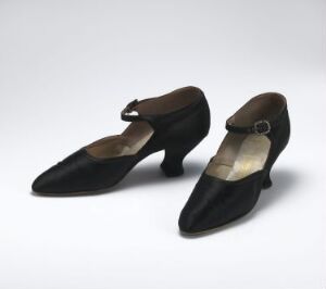  A pair of elegant vintage black pumps with pointed toes and low wide heels, featuring a small decorative strap over the instep, displayed against a neutral grey background.