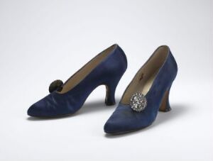  A pair of elegant deep blue high-heeled shoes with decorative elements, one with a blue bow and the other with a jewel brooch, set against a soft grey background.