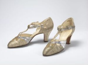  A pair of vintage champagne or light gold high-heeled T-strap women's shoes with a subtle shimmer, set against a neutral light grey background.