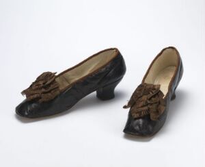  A pair of vintage black women's dress shoes with low, wide heels and decorative bows on the vamps, showing signs of wear, displayed against a light grey background.