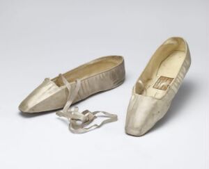  A pair of vintage, possibly satin, light-colored women's flats with pleated detailing, displaying one shoe with a detached decorative strap, against a light grey background.