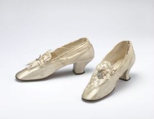  A pair of vintage ivory or light champagne-colored mid-heeled women's shoes with floral rhinestone embellishments on a neutral background. The shoes possess a subtle sheen and exhibit gentle wear, indicative of their historic charm and elegant design.