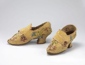  A pair of antique, creamy yellow women's shoes decorated with floral embroidery in hues of green, red, blue, and brown, featuring a low heel and pointed toes, displayed against a neutral gray background.