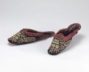  A pair of ornate, pointed traditional slippers with detailed gold and silver embroidery and colorful accents, set against a neutral gray background.