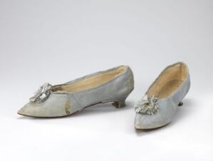  A pair of vintage pointed toe flats with low heels, in a subtle silvery-grey fabric with decorative bows and embellishments on the vamps, displayed against an off-white background.