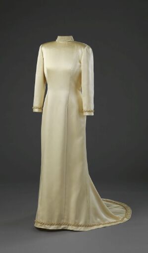  An elegant, long ivory dress with a high neckline, straight sleeves, and a trailing hem displayed against a dark gray backdrop, suggestive of classic formalwear.