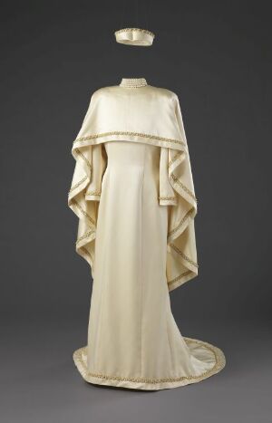  An elegant mannequin dressed in a historical-style cream gown with a structured cape overlay and a matching halo accessory, displayed against a dark grey background.