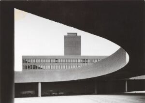  Black-and-white photograph of a modernist architectural structure with a curvilinear foreground framing a multi-story building with a tower in the background, showcasing a contrast of geometric shapes and an interplay of light and shadow.