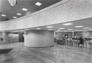  Black-and-white photograph of a mid-20th century modern interior architecture with a large semi-circular reception desk, square recessed ceiling lights, and two individuals. The space exhibits a minimalist design with a polished floor and textured surfaces.