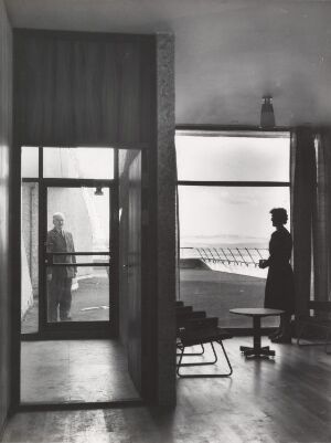  Black and white architectural photograph depicting an indoor scene with an open glass door and a woman in silhouette looking out at a seascape through a large window. The room features minimalistic design, highlighted by the interplay of light and shadow.
