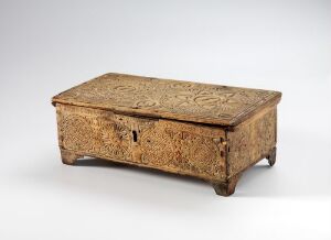  An antique, intricately carved wooden chest with a flat top, sitting on four short feet, showcased against a light gray background. The chest's varying brown tones and visible keyhole on the front panel suggest its age and use.