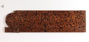  A long, horizontal, reddish-brown decorative panel with intricate scrolling vine and foliate motifs, suggesting a polished wooden material against a simple, neutral background. The piece exudes a sense of traditional craftsmanship, possibly used as architectural woodwork or furniture decoration.