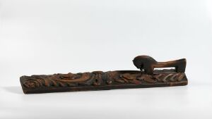  A dark brown, intricately carved wooden object with a flat base and a handle that curves upwards, resembling a bird's head, set against a plain, light background. The carving features a continuous, organic floral pattern along its length. Artist name and title are unknown.