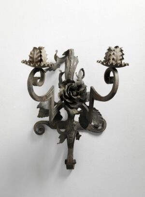  A wall-mounted candelabrum with a dark patina, featuring ornate swirling arms and a central rose design, intended to hold two candles against a plain, light-colored background.