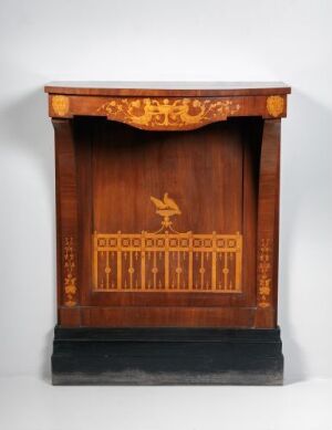  An antique wooden pulpit or lectern with dark mahogany tones and golden-hued inlay designs featuring a central bird in flight, sitting on a black base against a light grey backdrop.