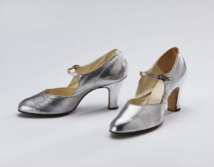  A pair of vintage-style silver women's shoes with modest heels and a strap across the instep, positioned against a white background.