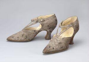  A pair of historical-style women's high-heeled shoes with pointed toes and a textured, mottled grey and brown surface. They feature T-straps with buttons and a contrasting light yellow interior lining.