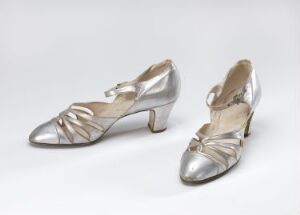  A pair of elegant metallic silver high-heeled shoes with crisscross straps and a light tan interior, set against a neutral gray background.