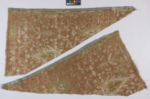  A torn fragment of an intricately patterned fabric in various shades of brown with floral designs, suggesting an antique textile piece, with a color chart in the upper left corner for reference.