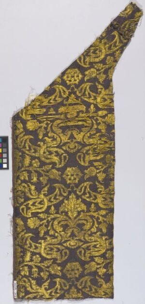  A fabric swatch with an irregular pentagonal shape, featuring an intricate golden-yellow pattern on a dark background, likely meant for textile analysis as evidenced by the inclusion of a ruler for scale.