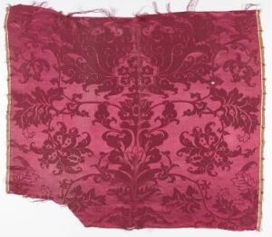  A piece of burgundy fabric with a symmetrical, faded pattern, featuring an elaborate central motif surrounded by intricate scrollwork and smaller floral designs, with frayed edges, indicative of a historical textile piece.