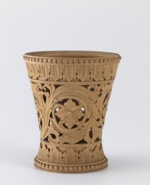  An intricately carved, cylindrical, light beige decorative object with openwork patterns, featuring a central floral medallion and surrounded by geometric designs, against a neutral grey background. The piece has a flared top edge and appears to be made of ceramic or carved stone.