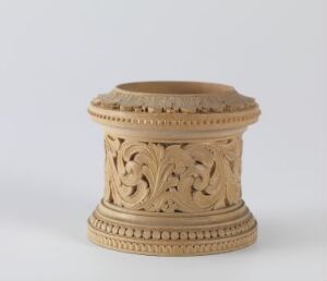  A cylindrical terracotta object with intricate leafy carvings in the center and dotted borders above and below, standing against a neutral grey background. Artist name and title are unknown.