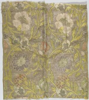  An old fabric featuring a symmetrical floral pattern with large blossoms in shades of pale cream and muted yellows, set against a grayish-beige background with details in olive green, possibly indicating a vintage textile with a fold or seam down the center.