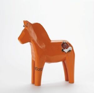  "Dalahest" by Katarina Brieditis – A glossy terracotta orange painted wooden Dala Horse with decorative black, white, green, and red folk art detailing on its body, standing against a neutral white background.