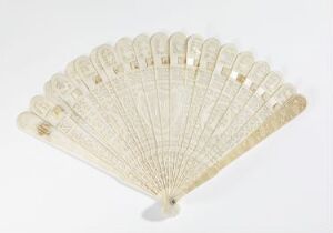  An elegantly crafted hand-held fan with creamy off-white sticks, each intricately carved or etched, fully opened against a light background. The fine, similarly colored material between the sticks is subtly embroidered, contributing to the fan's vintage charm.