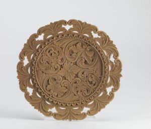  An ornate round wooden-like decorative object with a detailed, symmetrical floral and scroll design, set against a plain light background, suggestive of skilled craftsmanship. Artistname and title are unknown.