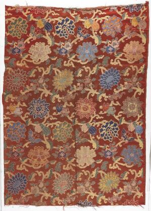  An antique-style textile featuring an intricate floral pattern with earthy brown background and motifs in shades of red, blue, yellow, and beige, suggesting a well-crafted, possibly hand-made rug or tapestry.